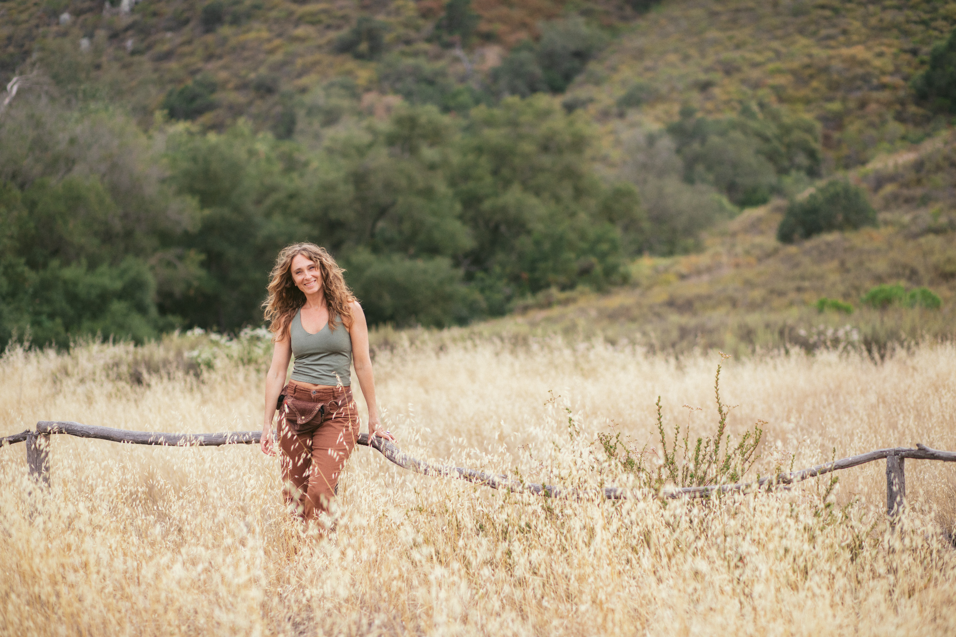 Jennica standing in a field of golden grass beside a weathered wooden fence, oak hills behind her