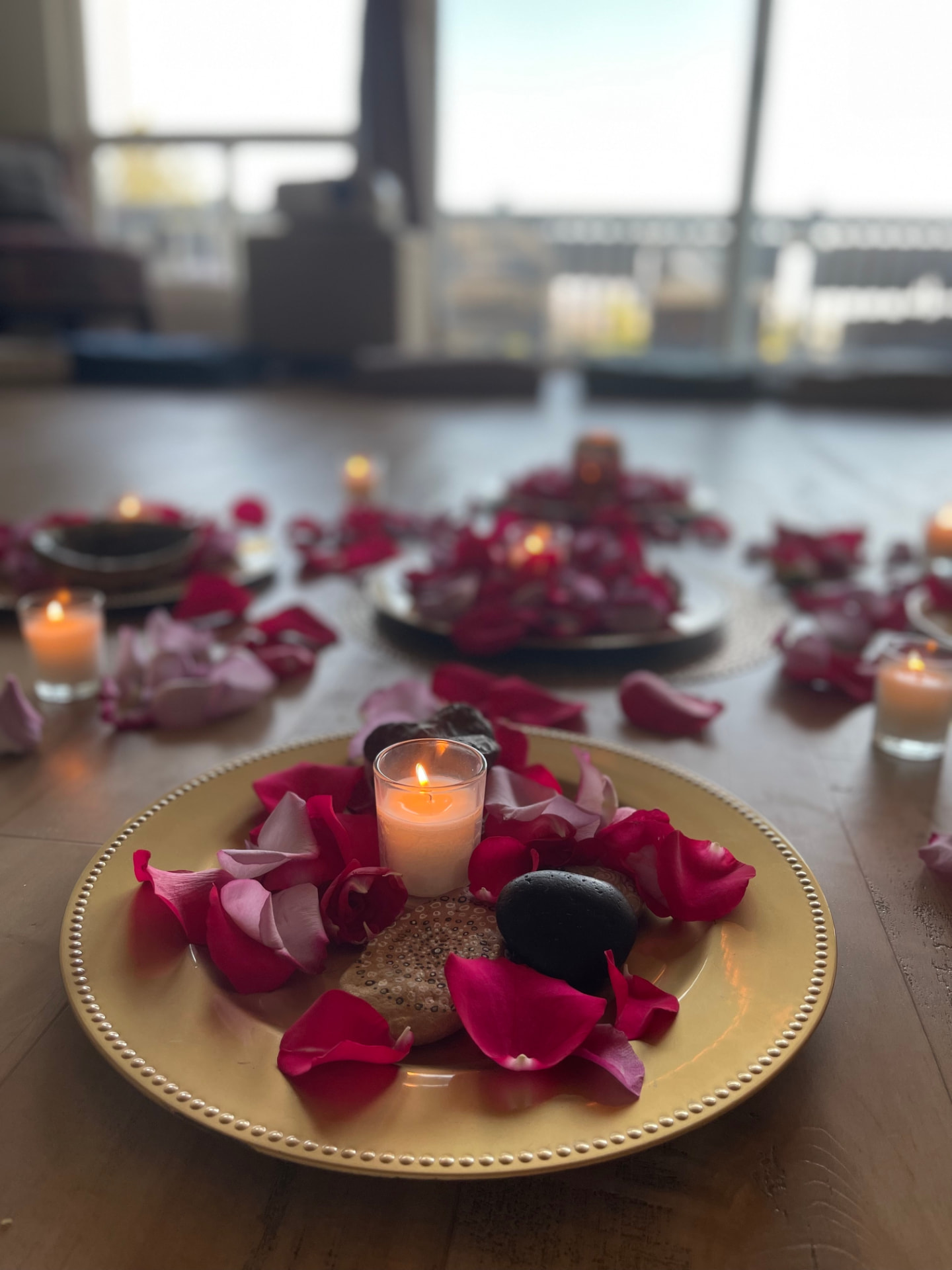 A candle surrounded by rose petals on a gold plate, with more petals and candles arranged across a wooden floor